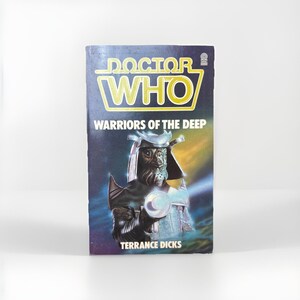 May include: A vintage paperback book titled "Doctor Who: Warriors of the Deep." The cover features a dark blue background with the title in yellow and white. An illustration of a creature with a helmet and glowing orb is in the center. The author's name, Terrance Dicks, is at the bottom.
