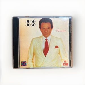 May include: A CD case featuring the album "Secretos" by José José. The cover art shows a man in a cream-colored suit and red tie. The album title and artist's name are displayed in a stylized font. The Ariola logo is visible.