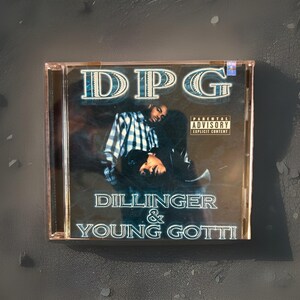 May include: A CD case featuring the album cover for "DPG - Dillinger & Young Gotti." The cover art shows two men, one in a plaid shirt. The album title and artist names are in a stylized font. The Parental Advisory label is visible.