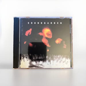 May include: A Soundgarden music CD in a clear plastic jewel case. The cover art features a dark, moody scene with glowing orange light shapes against a black background, positioned above a monochromatic, grainy image of a dense forest landscape.