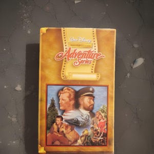 May include: A vintage Walt Disney Family Film Collection VHS tape from the Adventure Series. The cover features a collage of images, including portraits of men, a boy with a dog, and scenes of adventure. The title "Adventure Series" is prominently displayed.