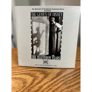 May include: A brochure for "The Genius of Disney: The Television Years" from The Museum of Broadcast Communications. The cover features a black and white image of a man and a silhouette of Mickey Mouse. The brochure is on a wooden surface.