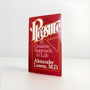 May include: A vintage paperback book titled "Pleasure: A Creative Approach to Life" by Alexander Lowen, M.D. The book has a red cover with white and gold lettering. The title is in a stylized font.