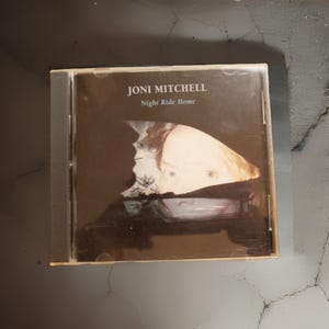 May include: A CD case featuring the album "Night Ride Home" by Joni Mitchell. The cover art displays a portrait with a dark background. The text "Joni Mitchell" and "Night Ride Home" are printed in white.