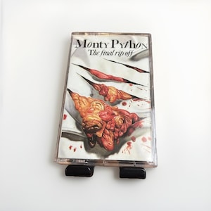 May include: A vintage cassette tape featuring the album art for "Monty Python: The Final Rip Off." The artwork depicts a torn surface with exposed flesh and blood, with the album title in black text.