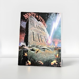 May include: A book cover for "Monty Python's The Meaning of Life" featuring a large stone with the title carved into it, set in a stormy sea. The cover art includes fish with human faces and lightning.