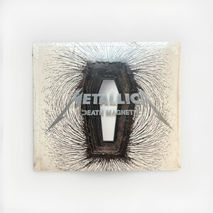 May include: A sealed album cover featuring the band name "METALLICA" and the album title "DEATH MAGNET". The cover art depicts a coffin shape with magnetic field lines radiating outwards. The design is in shades of black, silver, and white.