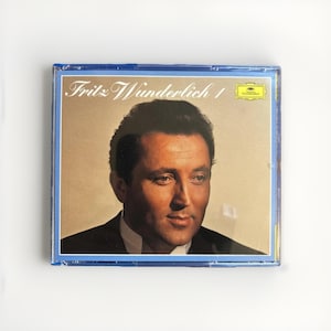 May include: A CD case featuring a portrait of a man in a suit. The CD case has a blue border and the text "Fritz Wunderlich 1" in a script font. The background is a light tan color.