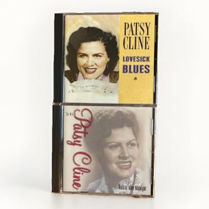 May include: Two stacked music CDs featuring Patsy Cline. The top case has a yellow cover with the text Patsy Cline Lovesick Blues. The bottom case has a white cover with the text The Best of Patsy Cline Walkin' After Midnight. Both show portraits.