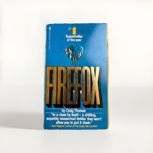 May include: A vintage paperback book titled "FIREFOX" by Craig Thomas. The cover is blue with gold lettering and features the text "Superthriller of the year." A quote from Jack Higgins is also visible. The book is in good condition.