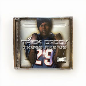 May include: A CD case featuring the album "Thugs Are Us" by Trick Daddy. The cover art shows a man in a blue jersey with the number 29. The album title and artist name are prominently displayed. The case has a parental advisory warning.