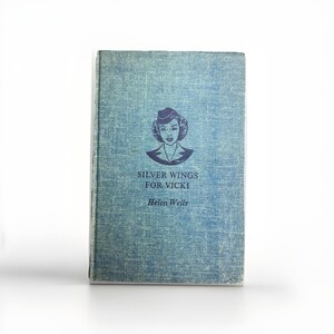 May include: A vintage book titled "Silver Wings for Vicki" by Helen Wells. The book has a blue cover with a graphic of a woman in a pilot's hat. The cover has a textured appearance.