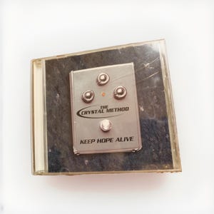 May include: A silver-colored effects pedal with four knobs and the text "THE CRYSTAL METHOD" and "KEEP HOPE ALIVE" printed on it. The pedal is housed in a clear plastic CD case.