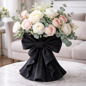May include: A floral arrangement featuring white and pink roses, eucalyptus, and other greenery. The flowers are arranged in a black velvet vase with a large bow detail. The vase sits on a marble-topped table.