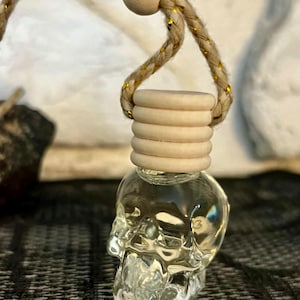 May include: A clear, skull-shaped glass bottle with a wooden cap and a braided rope. The rope is tan with gold accents and is attached to a wooden bead. The bottle is designed to hold liquid and is suspended by the rope.