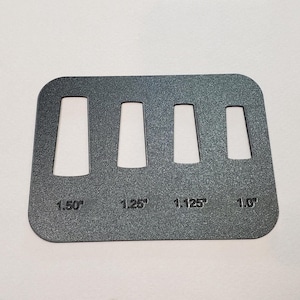 May include: A dark gray metal gauge with four rectangular cutouts of varying widths. The gauge is labeled with the measurements 1.50", 1.25", 1.125", and 1.0". The tool has rounded corners.