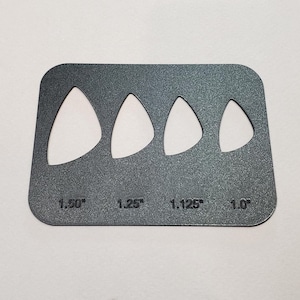 Peut inclure: Gabarit de médiator de guitare en métal gris foncé avec quatre découpes en forme de goutte d'eau de différentes tailles. Les tailles sont indiquées : 3,81 cm, 3,18 cm, 2,86 cm et 2,54 cm. Le gabarit a des coins arrondis.