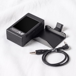 Op de afbeelding: Een zwart rechthoekig elektronisch apparaat met de tekst POWERPLAY op de voorzijde, vergezeld van een bijpassende zwarte kunststof montagebeugel en een zwarte audiokabel met een haakse 3,5 mm jack-aansluiting aan één uiteinde.