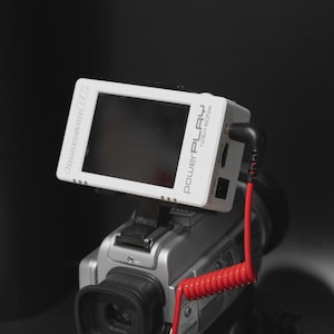 May include: A white PowerPlay video monitor attached to a silver video camera. The monitor has a black screen and the text "PowerPlay h264 60fps". A red coiled cable extends from the monitor.