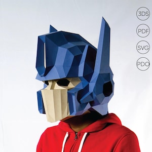 Op de afbeelding: Een blauw en beige papieren masker dat lijkt op een robothoofd, met hoekige vormen en details. Het masker wordt gedragen over een rode hoodie. De afbeelding bevat tekstlabels voor 3DS-, PDF-, SVG- en PDO-bestandsformaten.