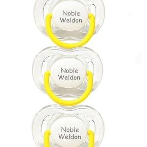 May include: Three clear baby pacifiers with yellow handles. Each pacifier has a white center with the text "Noble Weldon" printed in black. The pacifiers are arranged vertically against a white background.