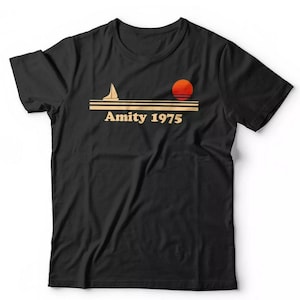 Op de afbeelding: Zwart t-shirt met een retro design met een zeilboot, een ondergaande zon en de tekst "Amity 1975" in een vintage lettertype. De afbeelding is in oranje en beige tinten. Het shirt heeft korte mouwen en een ronde hals.