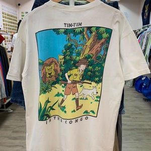May include: Cream-colored t-shirt featuring a colorful graphic from the Tintin comic book series, "Tintin in the Congo." The illustration depicts Tintin, a dog, a lion, and jungle foliage. The text "TIN-TIN" and "IN THE CONGO" are visible.