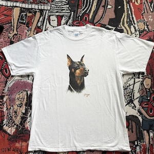 Könnte beinhalten: Weißes T-Shirt mit einem detaillierten Porträt eines Dobermanns. Der Kopf des Hundes ist in Braun-, Schwarz- und Beigetönen gehalten, mit einer Signatur in der unteren rechten Ecke. Das T-Shirt ist aus einem weichen, bequemen Material.