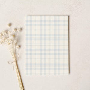 May include: A rectangular card with a light blue and cream plaid pattern. A bundle of dried, cream-colored flowers tied with twine is positioned to the left of the card, against a neutral background.
