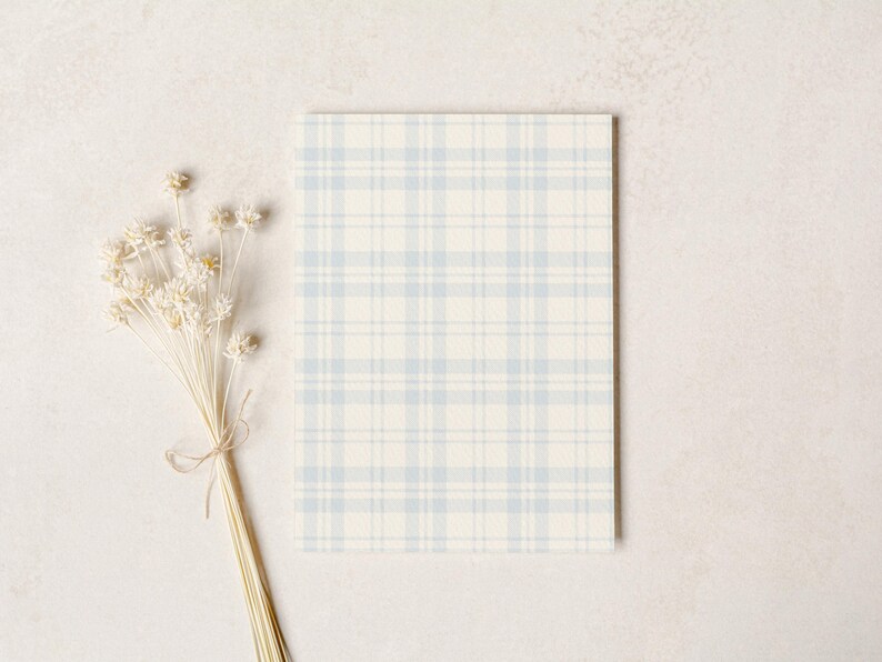 May include: A rectangular card with a light blue and cream plaid pattern. A bundle of dried, cream-colored flowers tied with twine is positioned to the left of the card, against a neutral background.