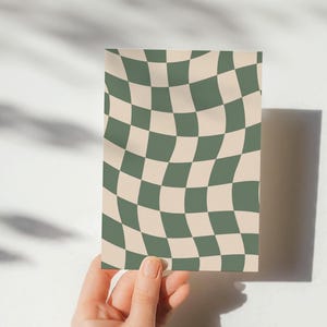 May include: A rectangular card with a wavy checkerboard pattern in olive green and beige. The card is held by a hand against a white background, with shadows cast on the surface.