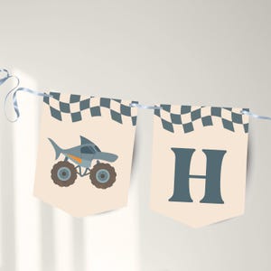 May include: A banner with two pennants. One pennant features a shark-shaped monster truck with orange accents and brown wheels. The other pennant displays the letter "H" in a teal color. Both pennants have a checkered flag design at the top.