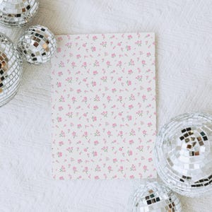 May include: A rectangular card with a white background is covered in a pattern of small pink flowers and green stems. Four disco balls with mirrored surfaces surround the card, creating a festive and decorative scene.
