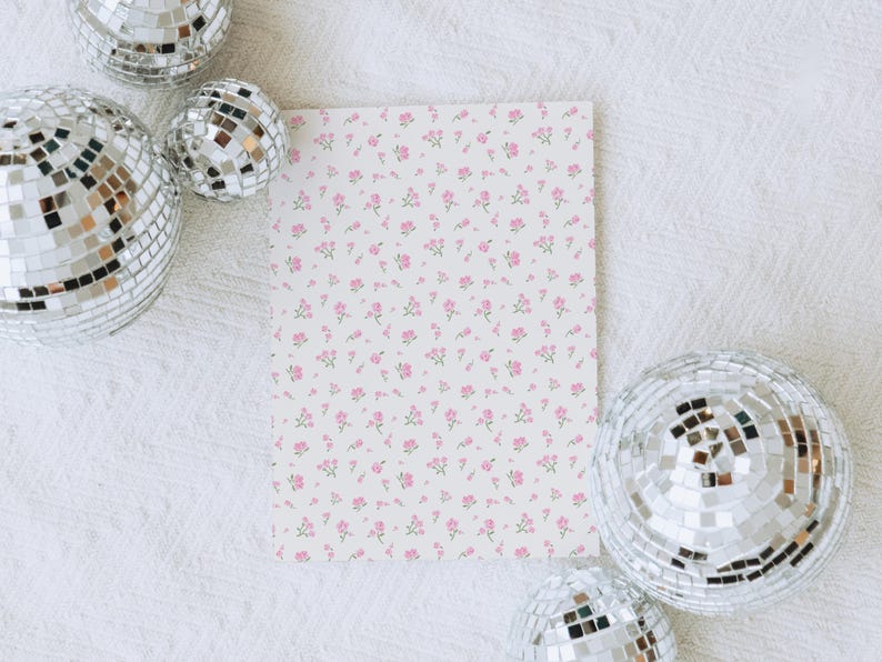 May include: A rectangular card with a white background is covered in a pattern of small pink flowers and green stems. Four disco balls with mirrored surfaces surround the card, creating a festive and decorative scene.