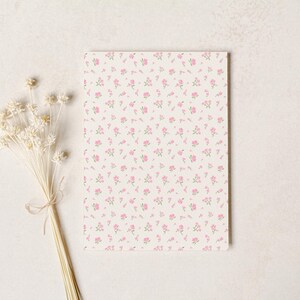 May include: A rectangular notepad with a cream background covered in small pink floral patterns. A bundle of dried white flowers tied with twine rests to the left of the notepad on a white surface.