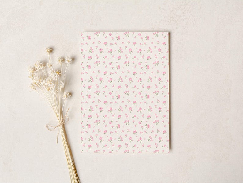 May include: A rectangular notepad with a cream background covered in small pink floral patterns. A bundle of dried white flowers tied with twine rests to the left of the notepad on a white surface.