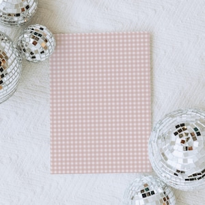 May include: A square, light pink and white gingham pattern card is surrounded by several silver disco balls on a white textured surface. The disco balls reflect light, creating a sparkling effect.