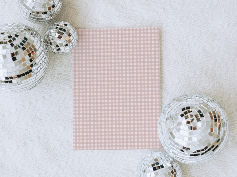May include: A square, light pink and white gingham pattern card is surrounded by several silver disco balls on a white textured surface. The disco balls reflect light, creating a sparkling effect.