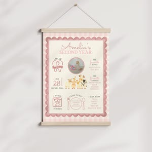 May include: A pink and cream-colored poster with the text "Amelia's Second Year." The poster includes illustrations of teeth, a baby, farm animals, and text indicating height at 28 inches and weight at 25 pounds. It is hanging from a wooden bar.