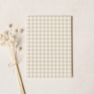 May include: A rectangular notebook with a light green and white gingham pattern. A bundle of dried, cream-colored flowers tied with twine rests to the left of the notebook on a neutral background.