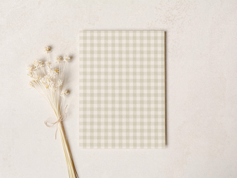 May include: A rectangular notebook with a light green and white gingham pattern. A bundle of dried, cream-colored flowers tied with twine rests to the left of the notebook on a neutral background.