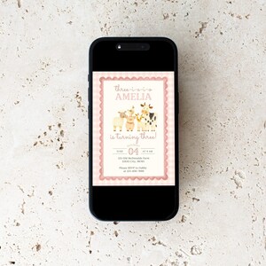 May include: A smartphone displays a pink and cream birthday invitation. The invitation features illustrations of farm animals and the text "three-o-two Amelia is turning three!" The date is June 4th.