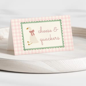 May include: A folded card with a pink and white gingham pattern. The card features a cartoon goose wearing a pink bow and the words "cheese & quackers". The card is on a white wooden tray.