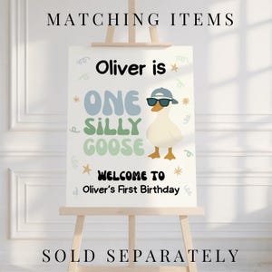 May include: A white sign on a wooden easel with the text "MATCHING ITEMS" at the top and "SOLD SEPARATELY" at the bottom. The sign reads "Oliver is ONE SILLY GOOSE" with a cartoon goose wearing sunglasses and a hat. The sign says "WELCOME TO Oliver's First Birthday."