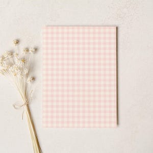 Puede incluir: Una tarjeta rectangular con un patr&oacute;n de cuadros rosa y blanco. Un ramo de flores secas de color crema, atadas con hilo, descansa a la izquierda de la tarjeta. El fondo es una superficie clara y texturizada.