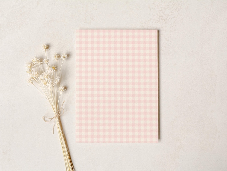 Puede incluir: Una tarjeta rectangular con un patr&oacute;n de cuadros rosa y blanco. Un ramo de flores secas de color crema, atadas con hilo, descansa a la izquierda de la tarjeta. El fondo es una superficie clara y texturizada.