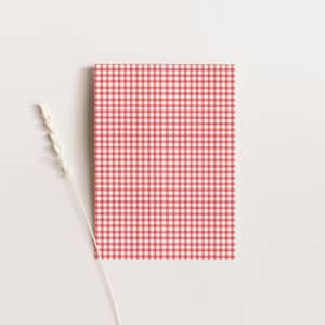 Pu&ograve; includere: Un taccuino rettangolare con un motivo a quadretti vichy rosso e bianco. Uno stelo di pianta beige secco &egrave; posizionato a sinistra del taccuino su uno sfondo bianco. Il taccuino ha un design classico.