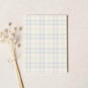 Op de afbeelding: Een rechthoekig notitieblok met een lichtblauw en wit ruitpatroon. Een bosje gedroogde, cr&egrave;mekleurige bloemen, vastgebonden met touw, is links van het notitieblok geplaatst. De achtergrond is een neutrale, gebroken witte kleur.