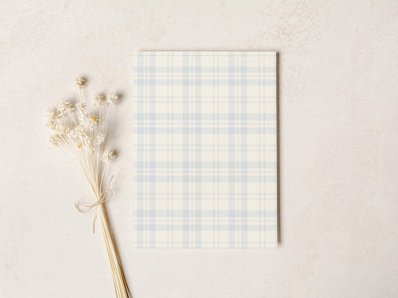 Op de afbeelding: Een rechthoekig notitieblok met een lichtblauw en wit ruitpatroon. Een bosje gedroogde, cr&egrave;mekleurige bloemen, vastgebonden met touw, is links van het notitieblok geplaatst. De achtergrond is een neutrale, gebroken witte kleur.