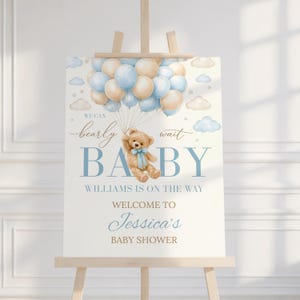 May include: A baby shower sign on an easel. The sign features a teddy bear holding balloons in shades of blue and tan. Text includes "We can bearly wait BABY" and "Welcome to Jessica's Baby Shower."